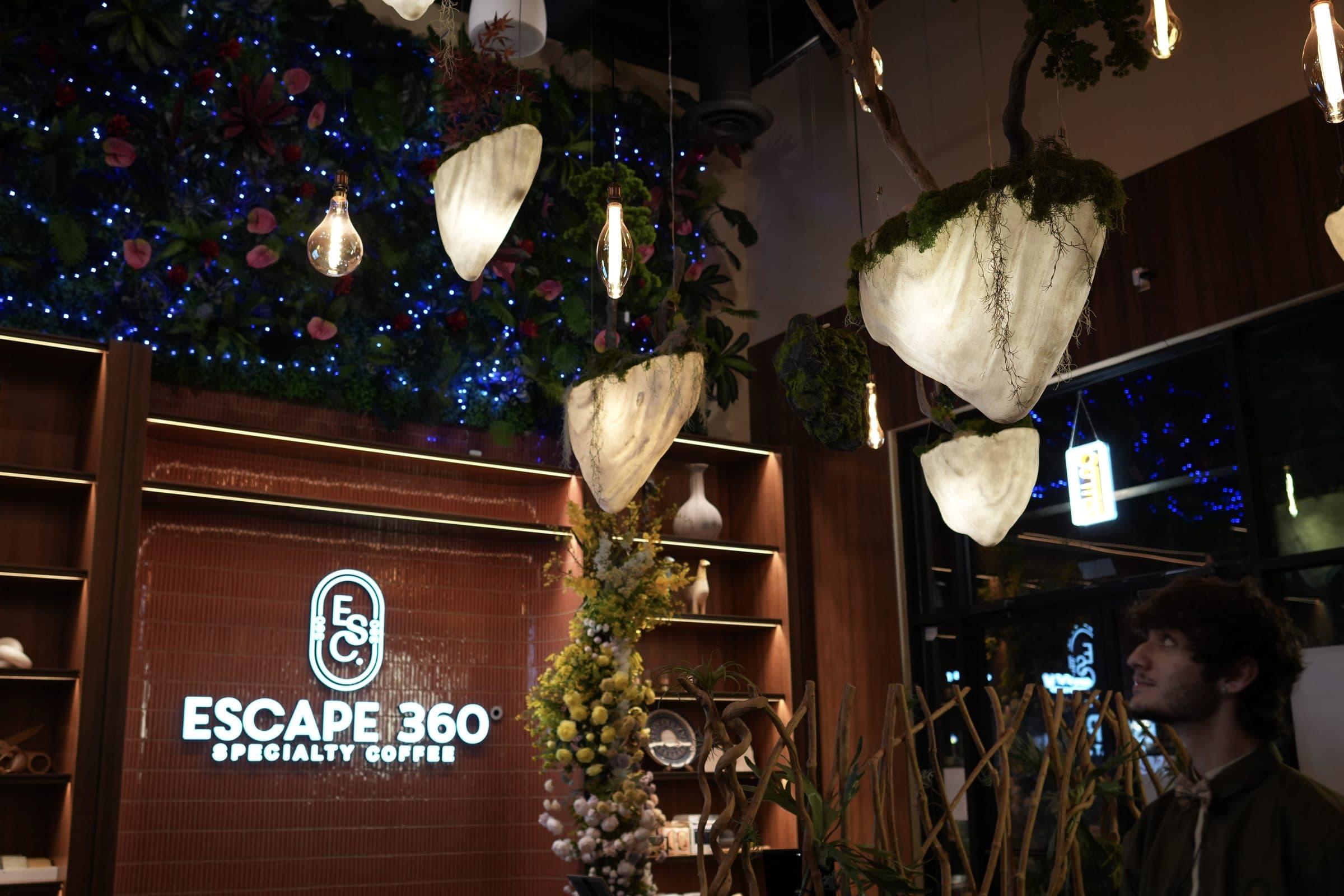 Floating moss-pod lanterns glowing over the flower wall and Escape 360 sign at the Carrollton cafe